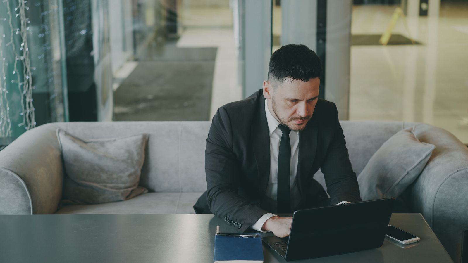 Man in a dark suit working on a laptop at a low table in an office lounge