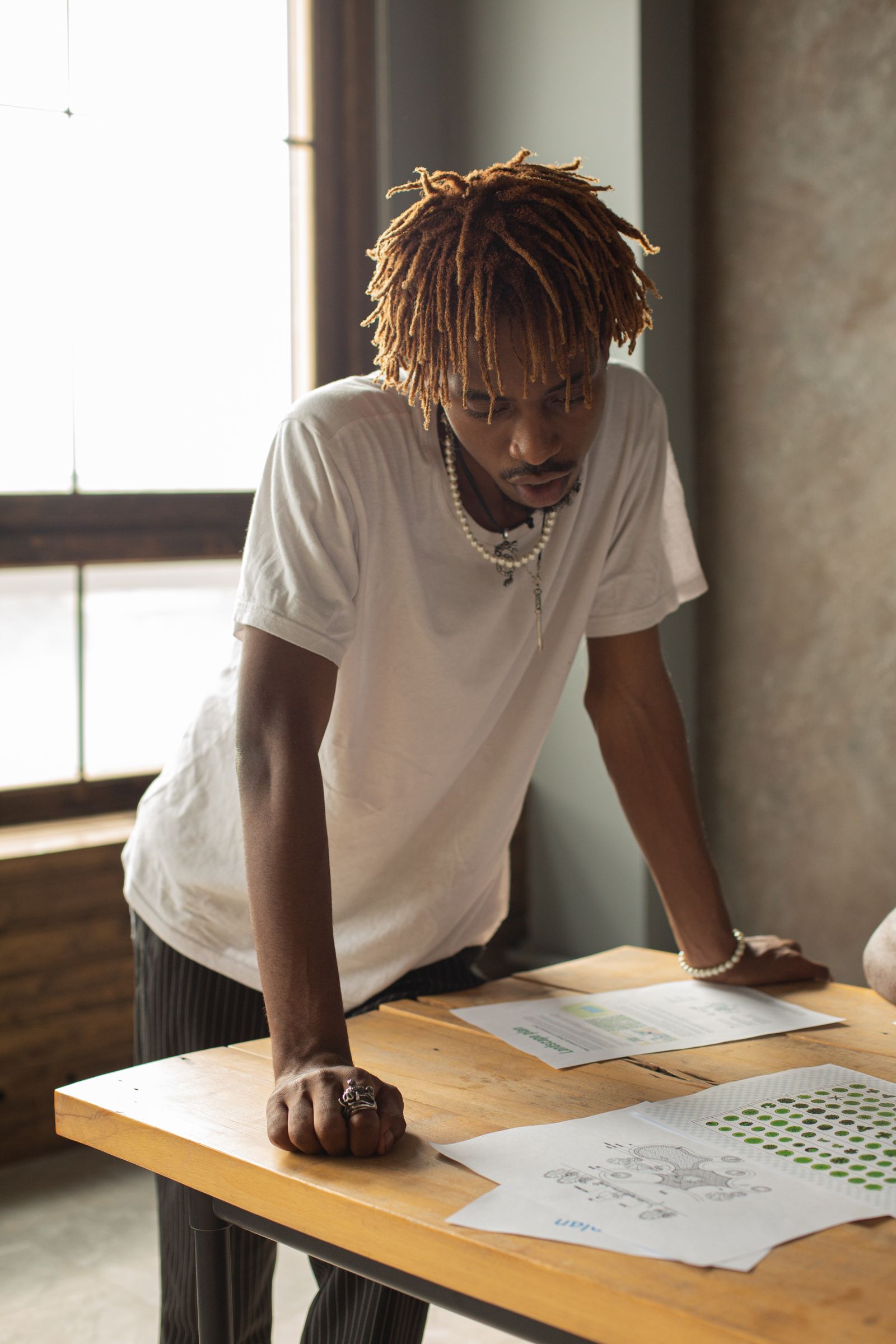 A man in a white T-shirt standing at a wooden desk reading papers by a window