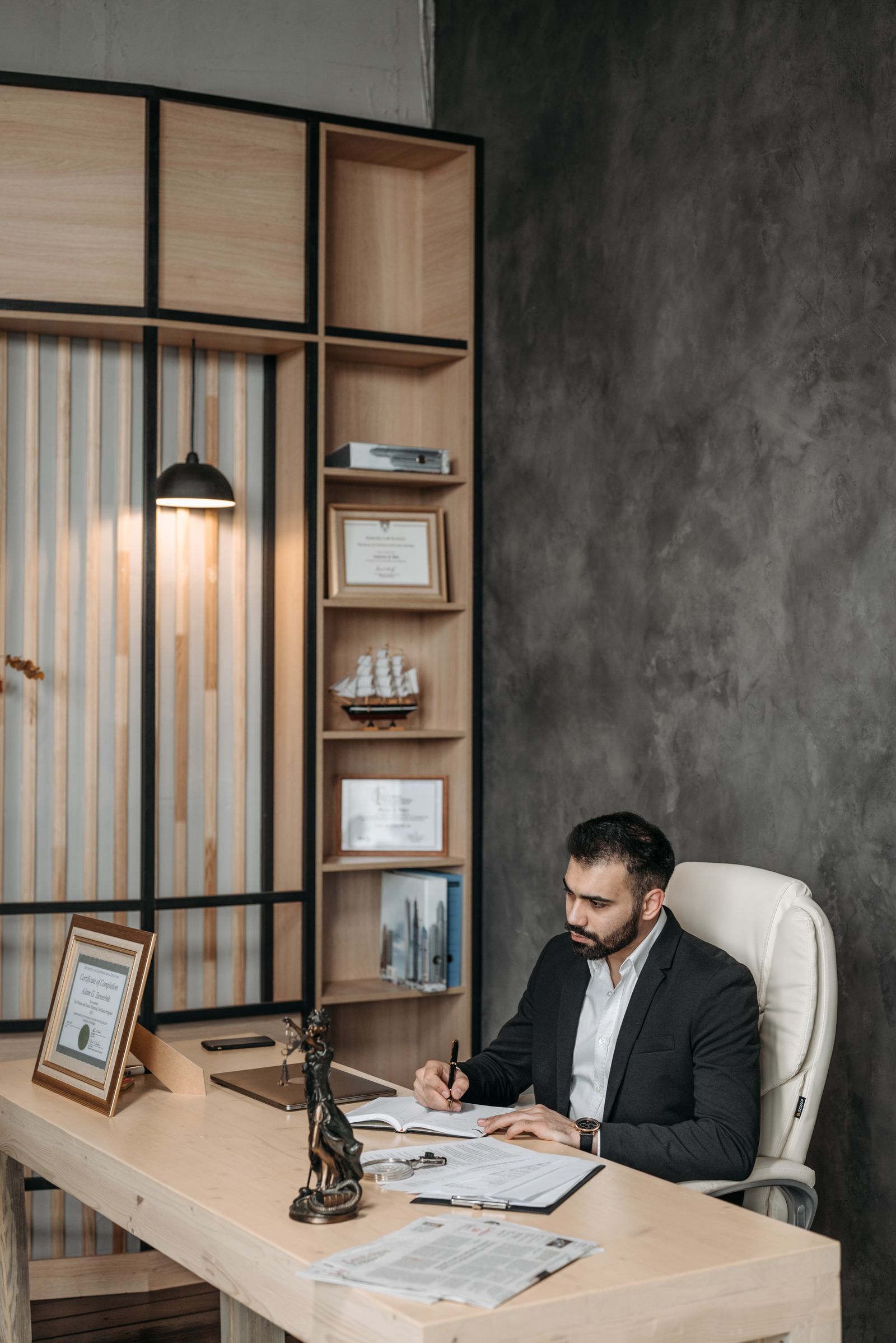 Man in a white shirt and dark jacket sitting at a wooden desk writing in a notebook