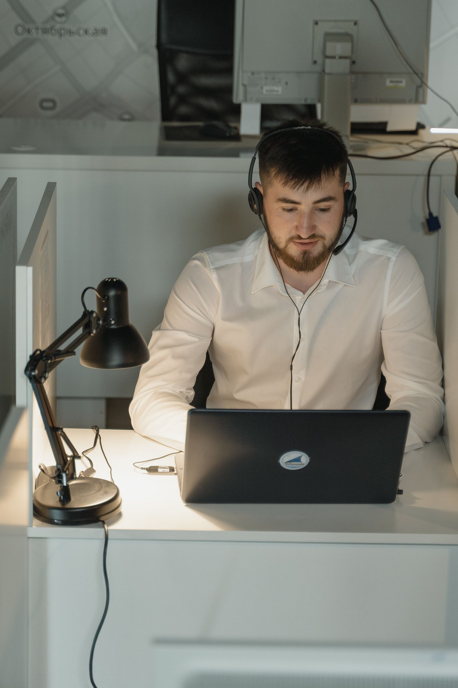 Man in a white shirt wearing a headset while working at a laptop in a desk booth