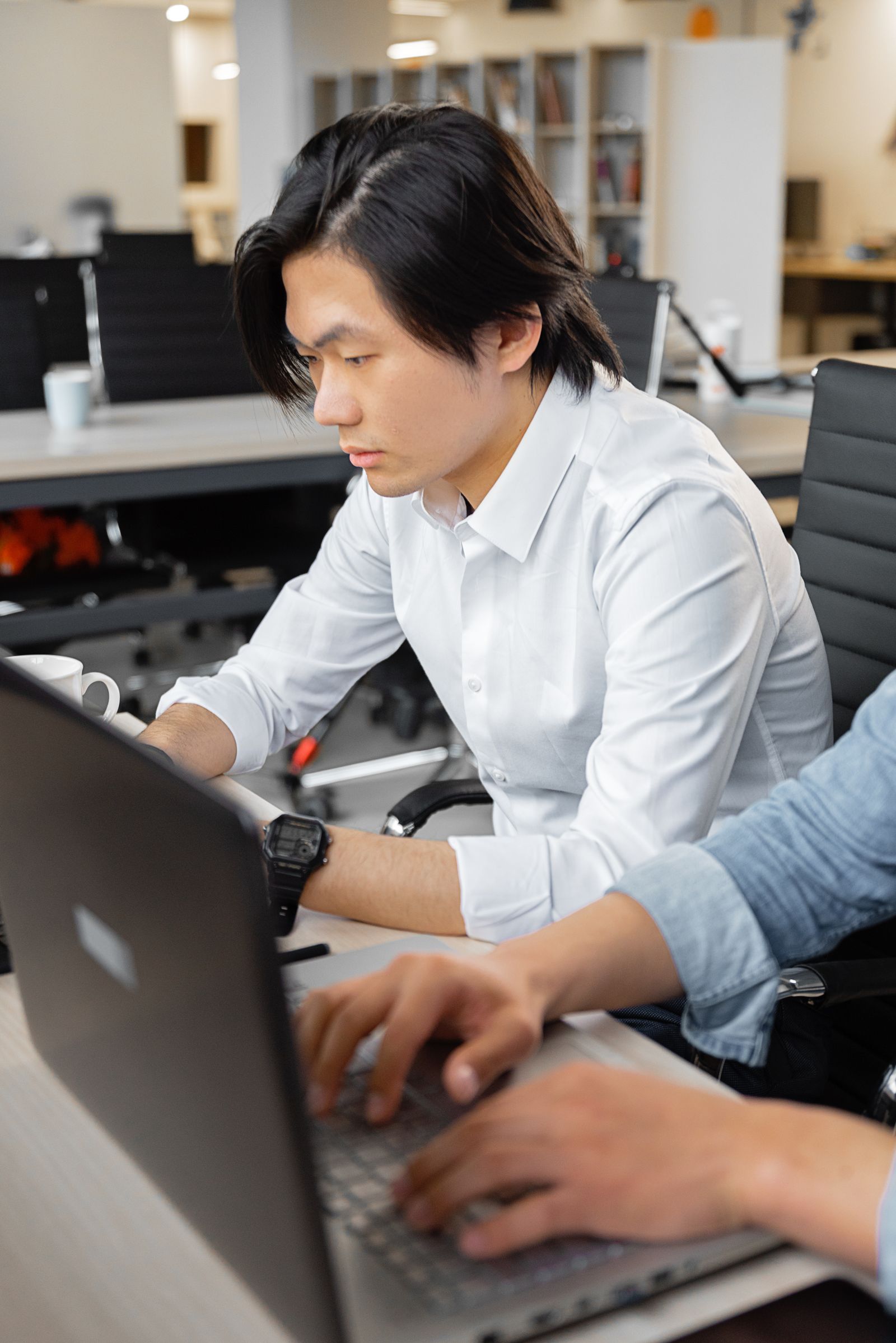 Man in a white shirt working on a laptop at a shared office desk beside a colleague