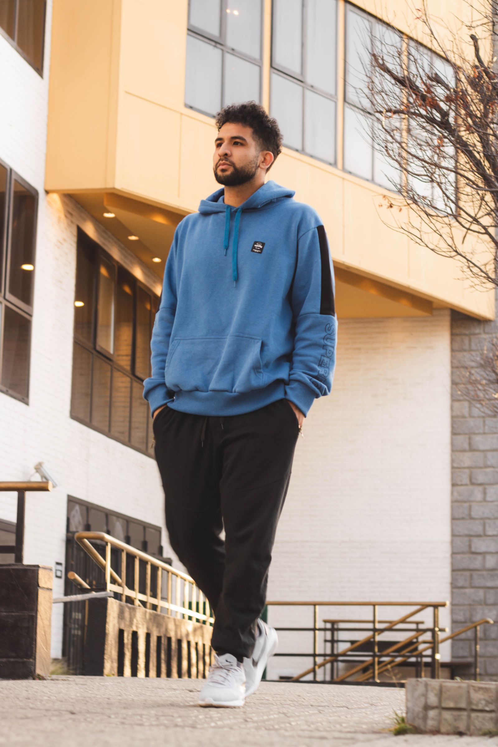 A man in a blue hoodie and dark trousers walking on a city pavement