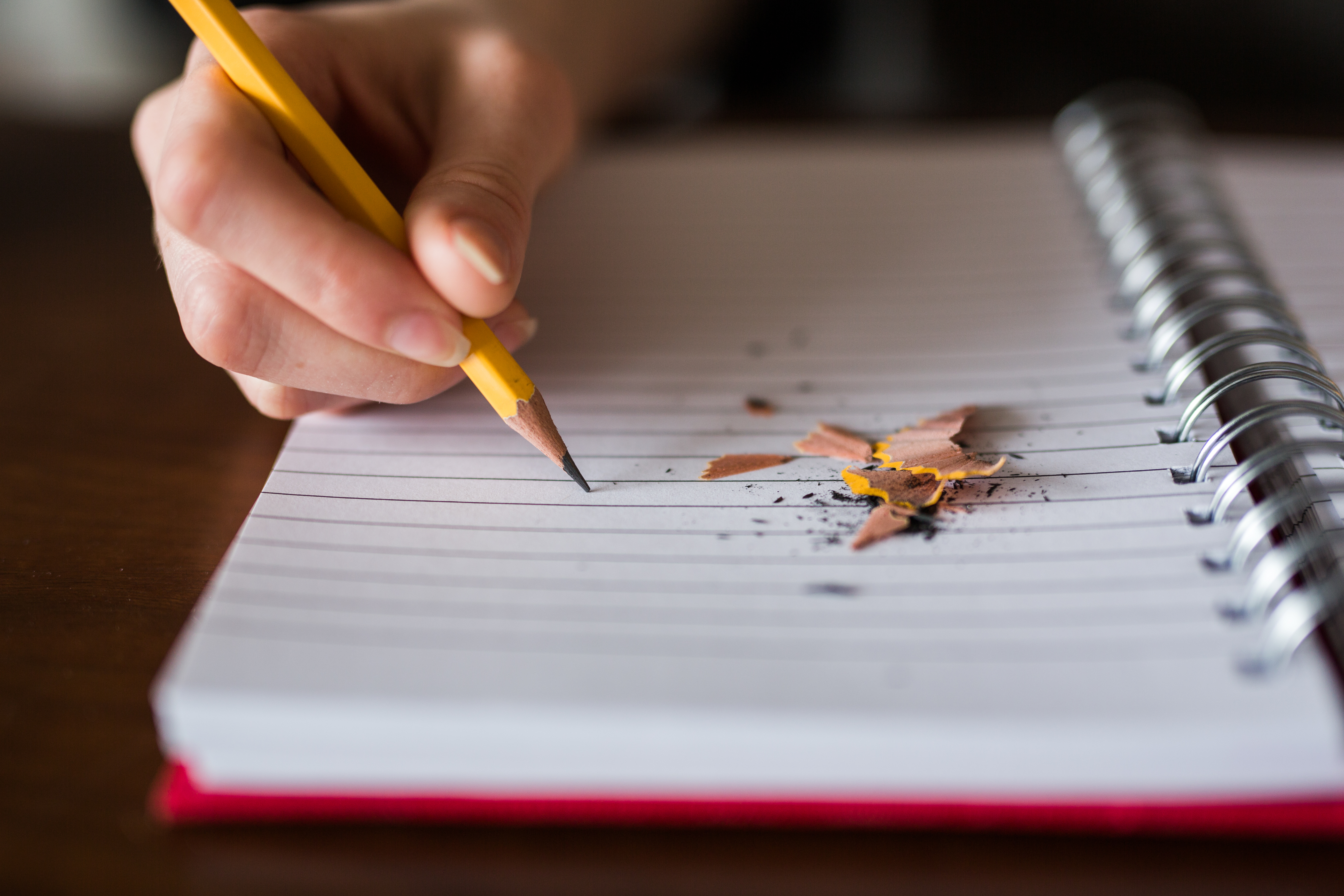 A hand holding a yellow pencil above a lined notebook with pencil shavings on the page