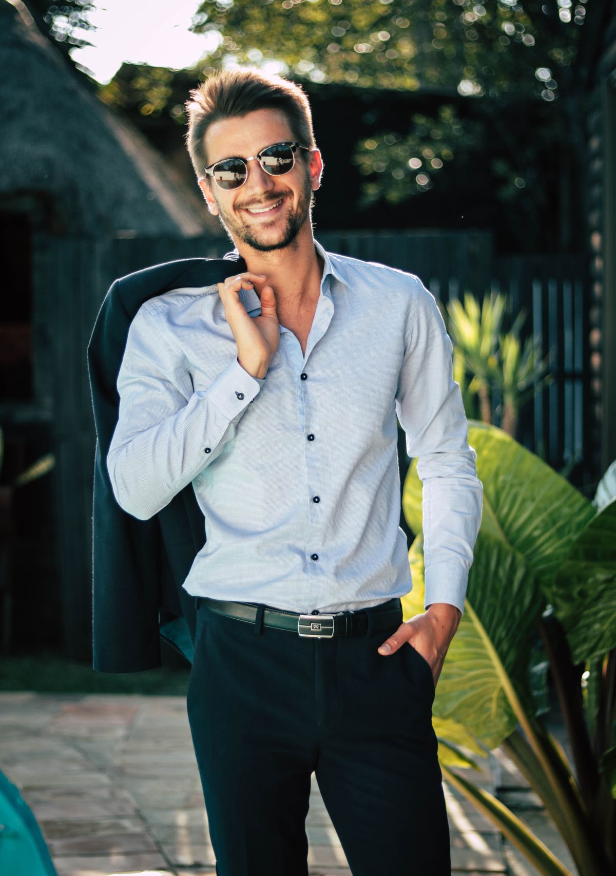 A smiling man in a light shirt and dark trousers standing outdoors in everyday clothes