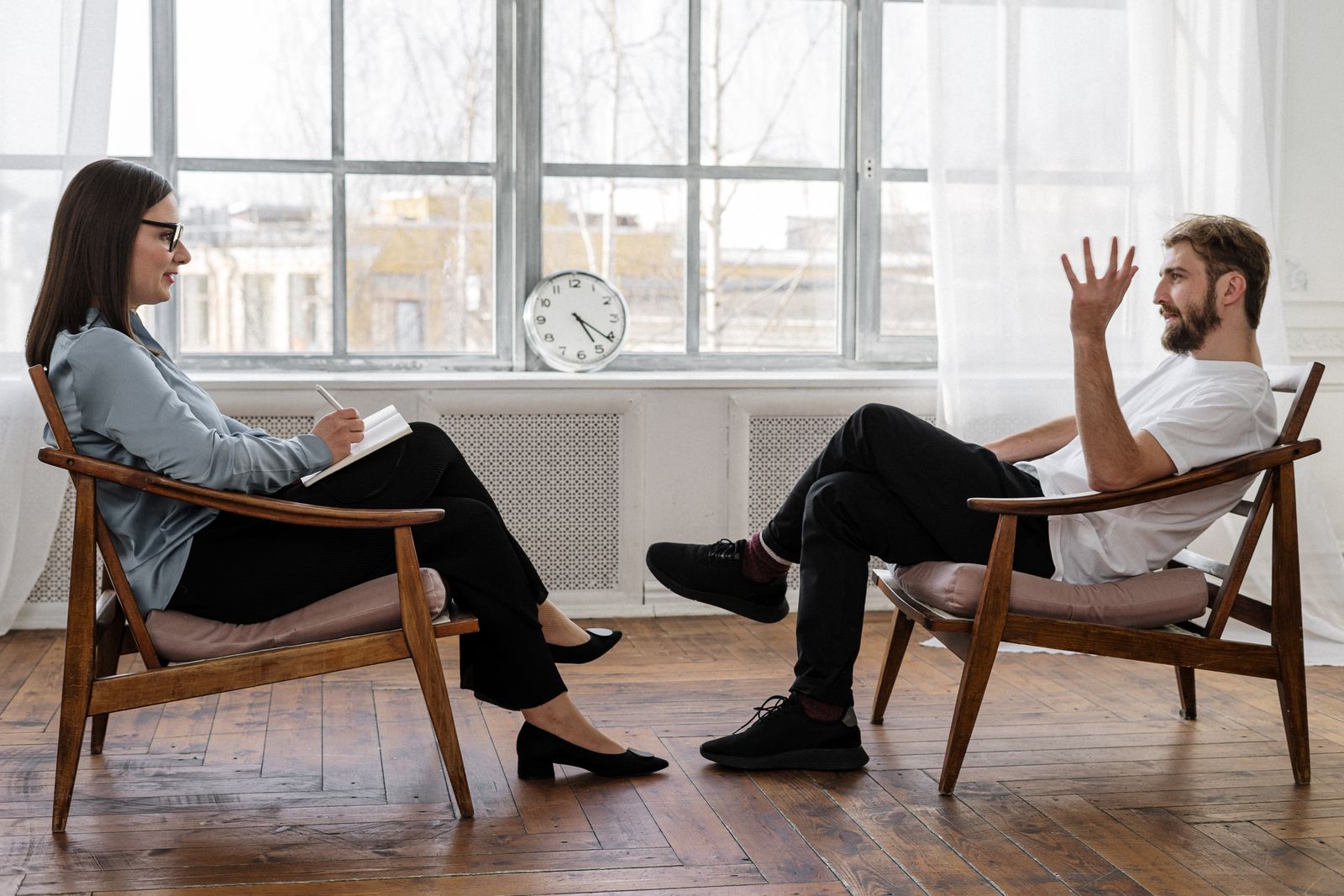 A man in a white t-shirt talking with a clinician who is taking notes by a window