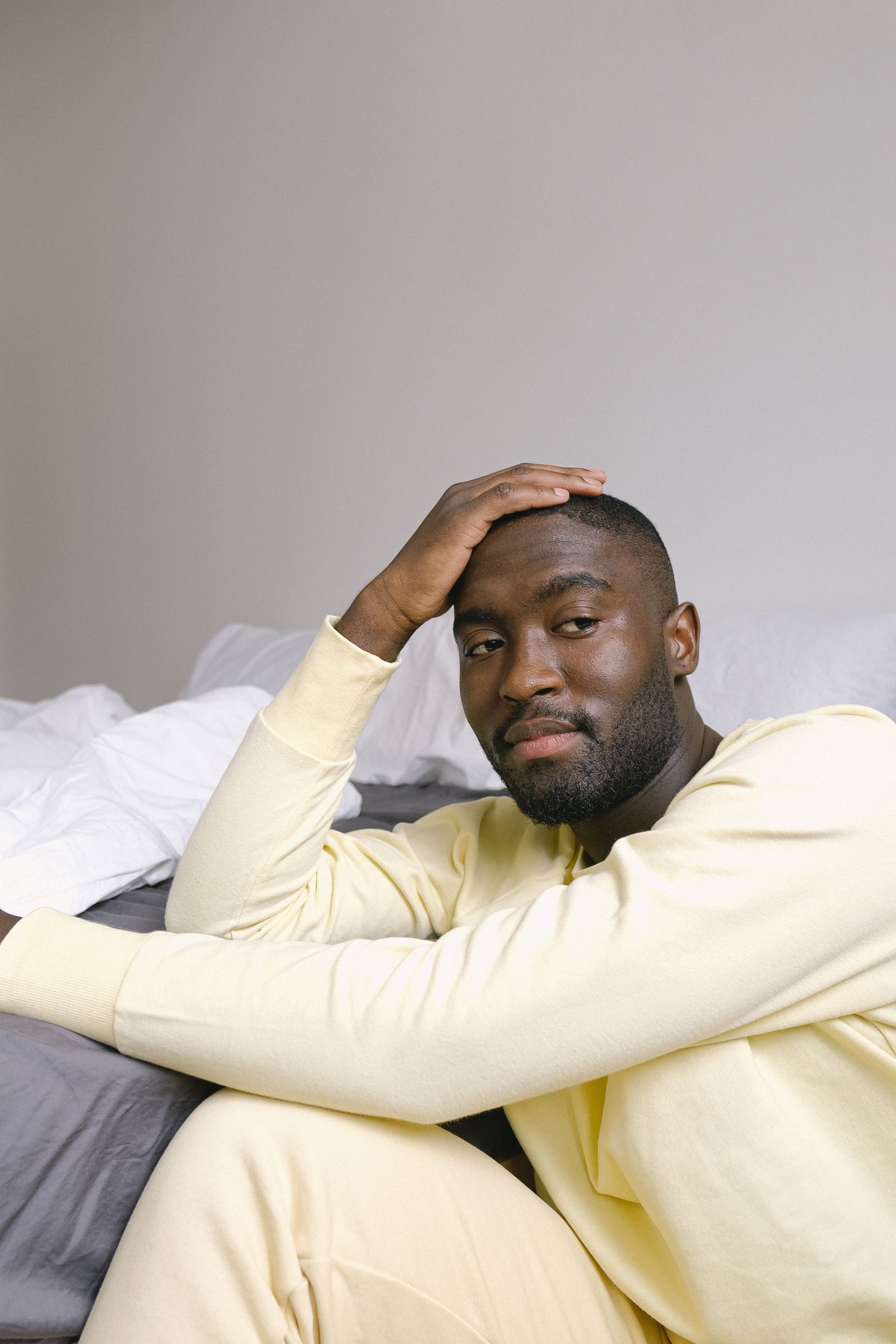 A man in a yellow long-sleeve top sitting on the floor beside a rumpled bed