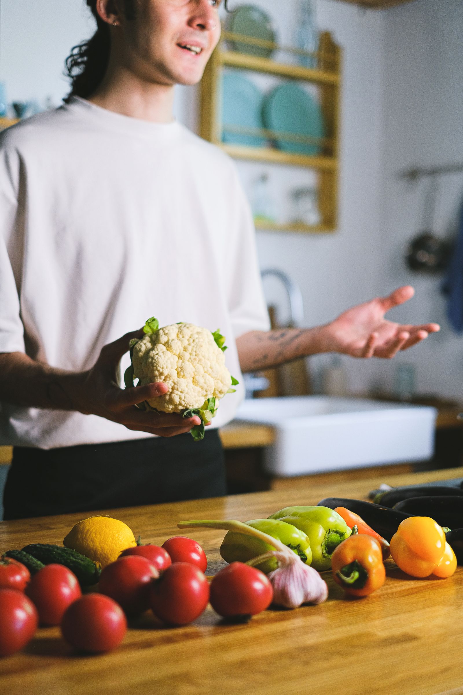 A man in a white T-shirt holding a cauliflower beside tomatoes and peppers on a kitchen counter