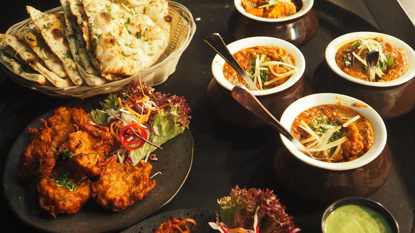 Shared Indian restaurant dishes with naan, curries in bowls, and a side salad on a dark table