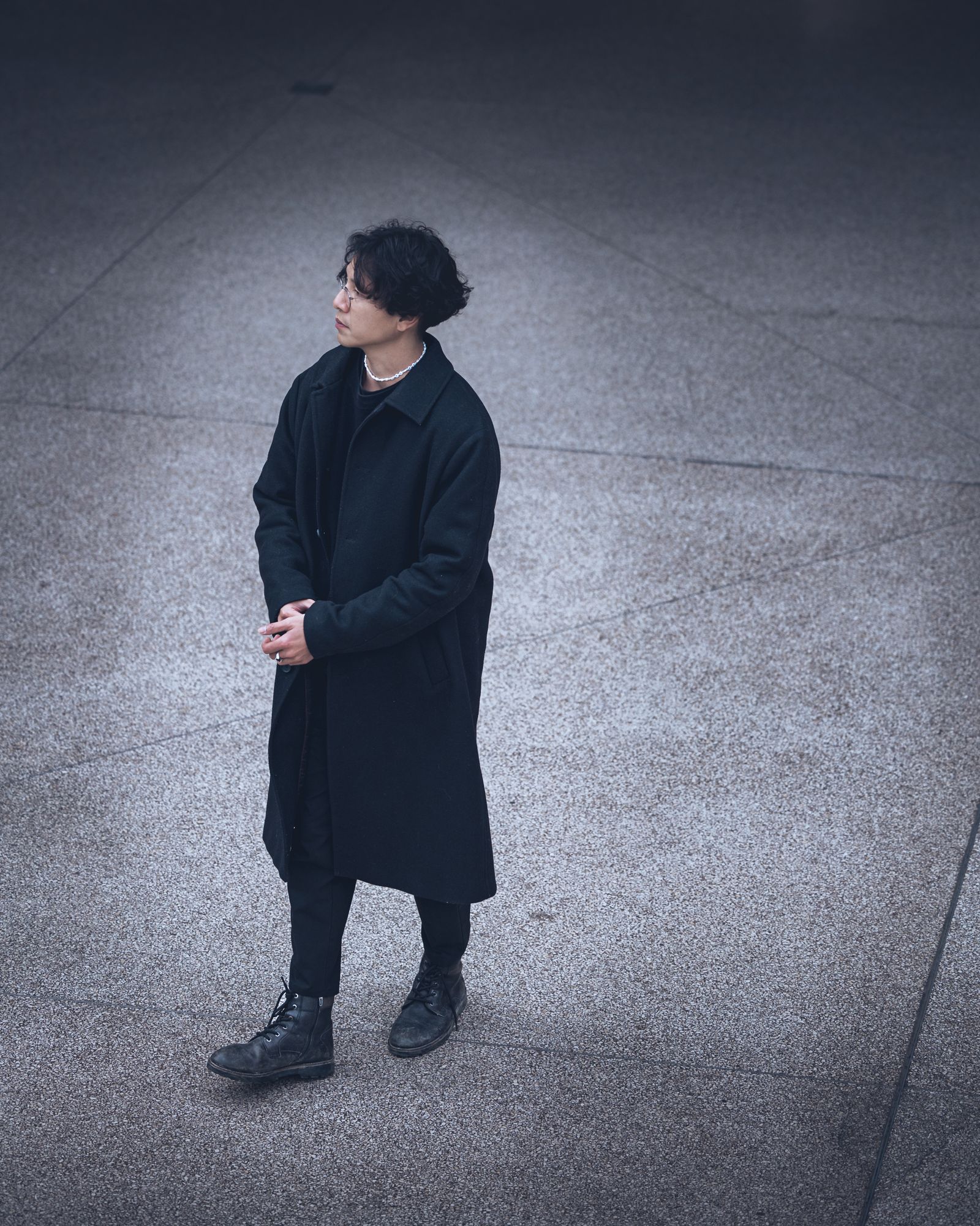 A man in a long dark coat and boots standing on a paved square in daylight