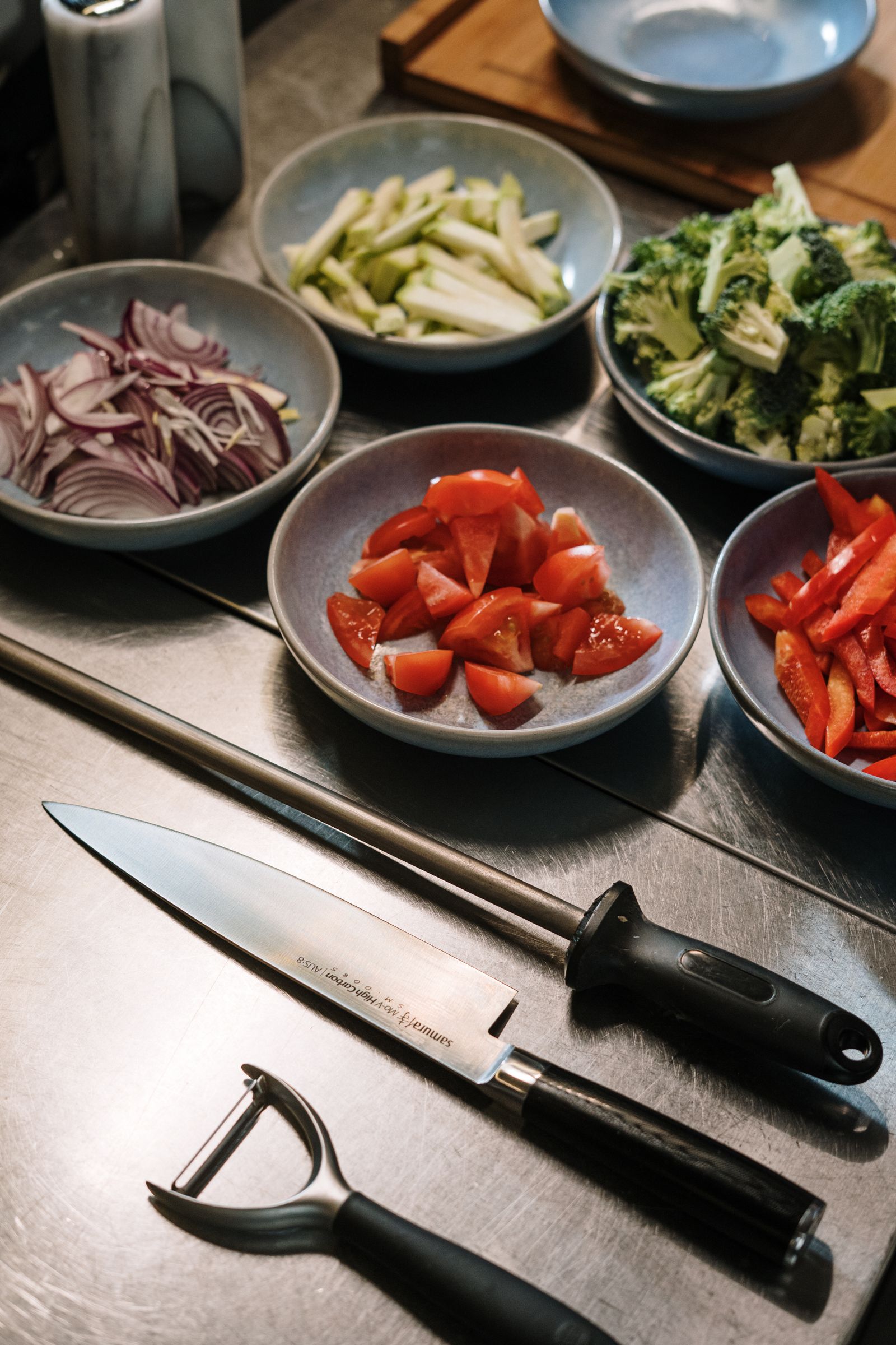 Bowls of chopped tomatoes, peppers, broccoli and onions beside a cook’s knife on a steel counter