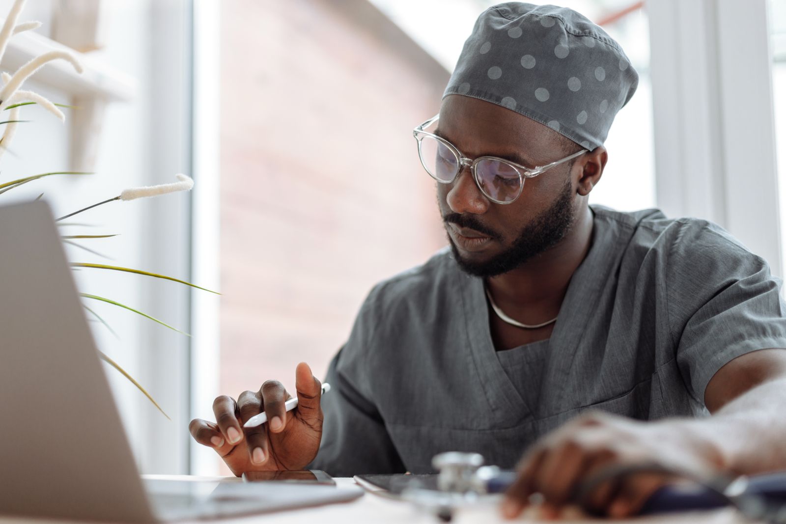 A clinician in grey scrubs and a cap reviewing notes on a laptop at a desk