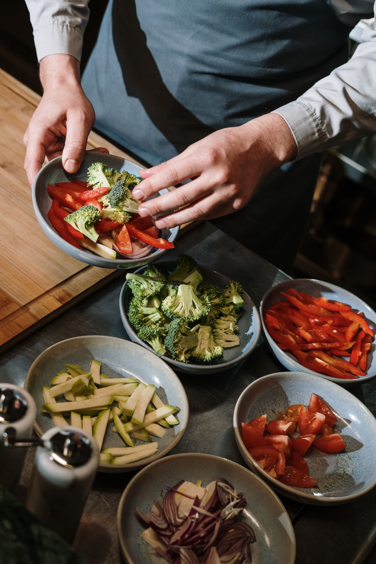 A cook in a shirt and apron arranging bowls of chopped peppers, broccoli and tomato