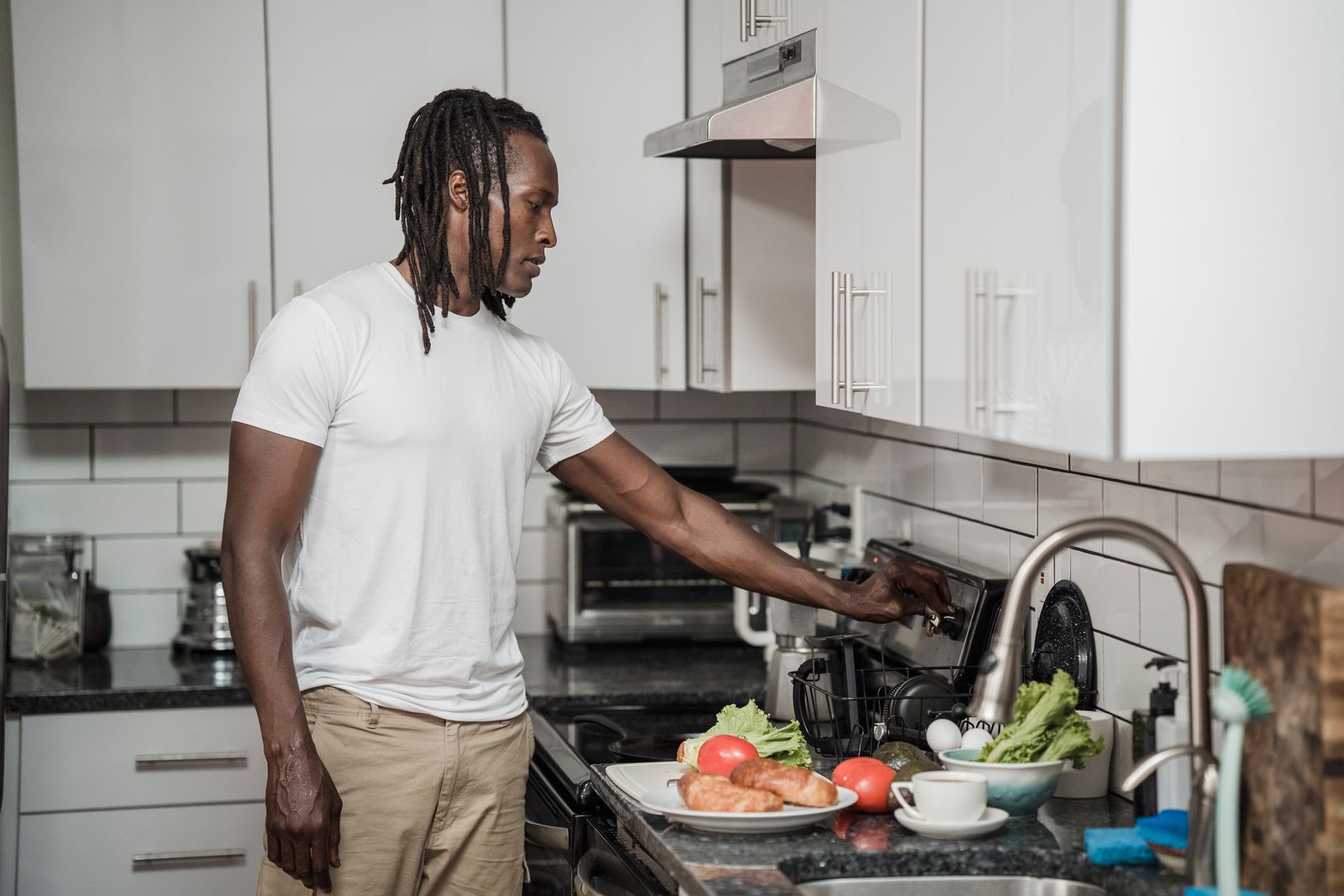 A man in a white t-shirt and beige trousers cooking breakfast at a home hob