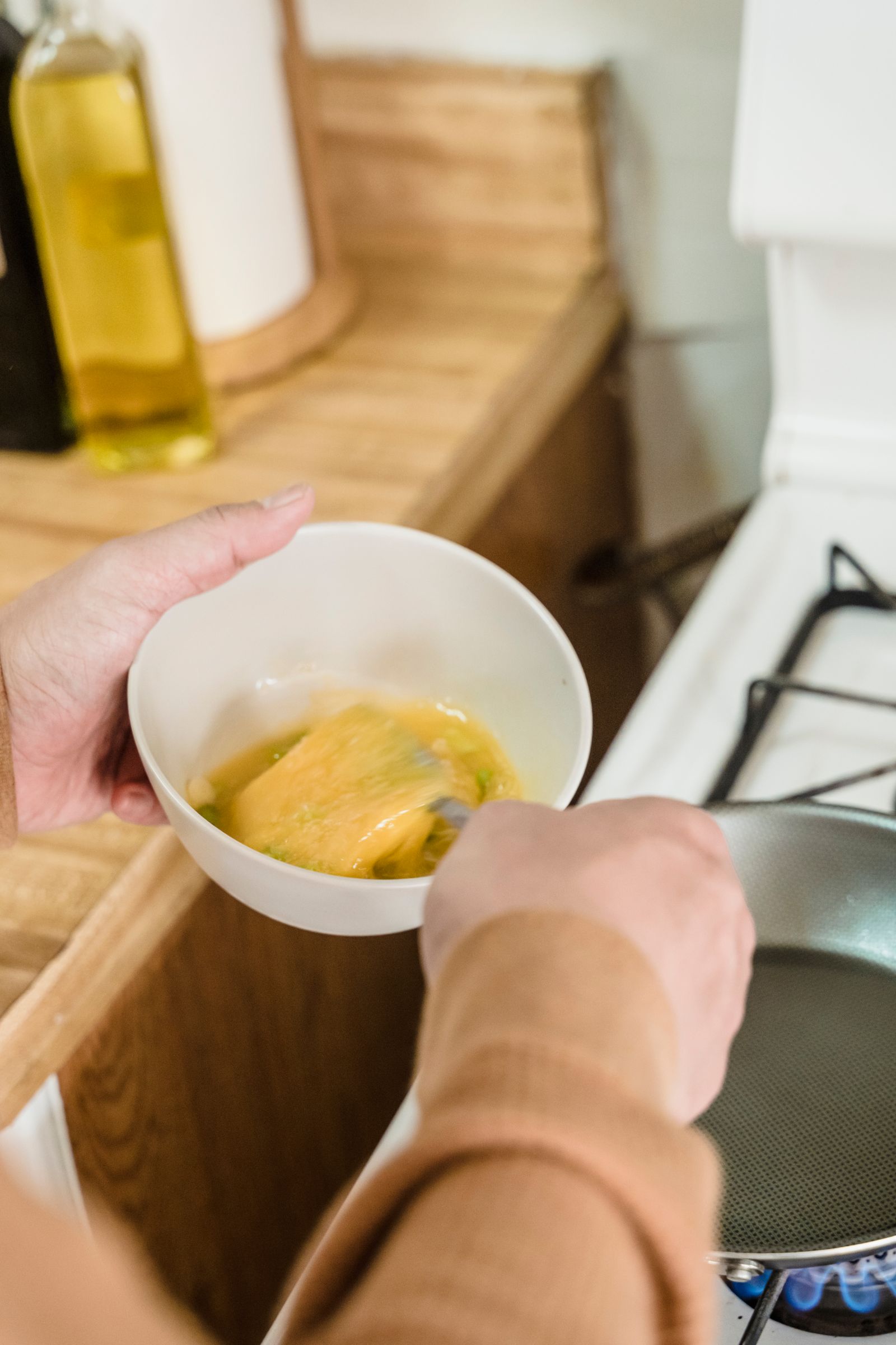 A person in a brown jumper whisking beaten eggs beside a pan on a home gas hob