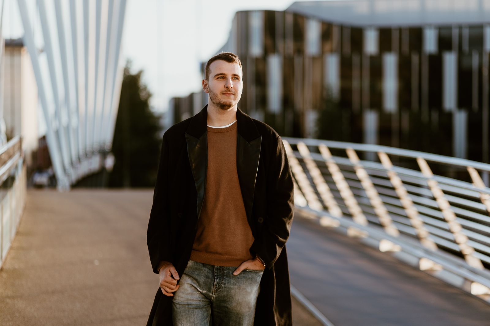 A man in a brown jumper and dark coat walking on a city footbridge in daylight