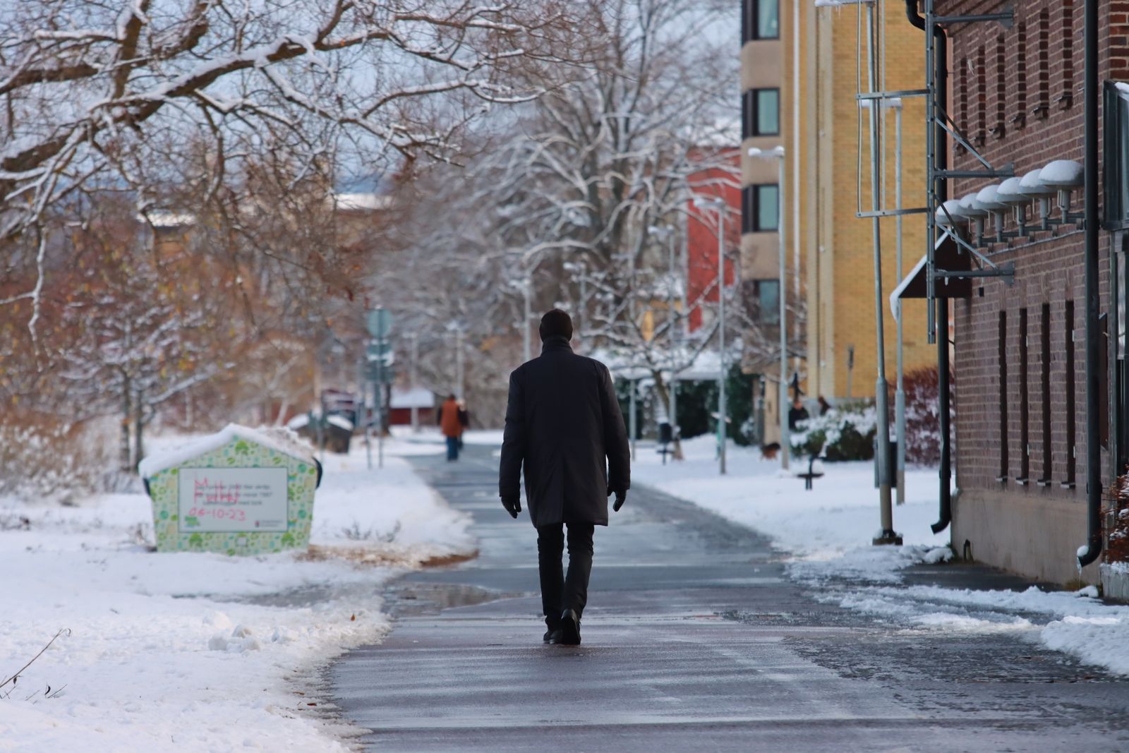 A man in a dark winter coat walking away along a quiet snowy pavement