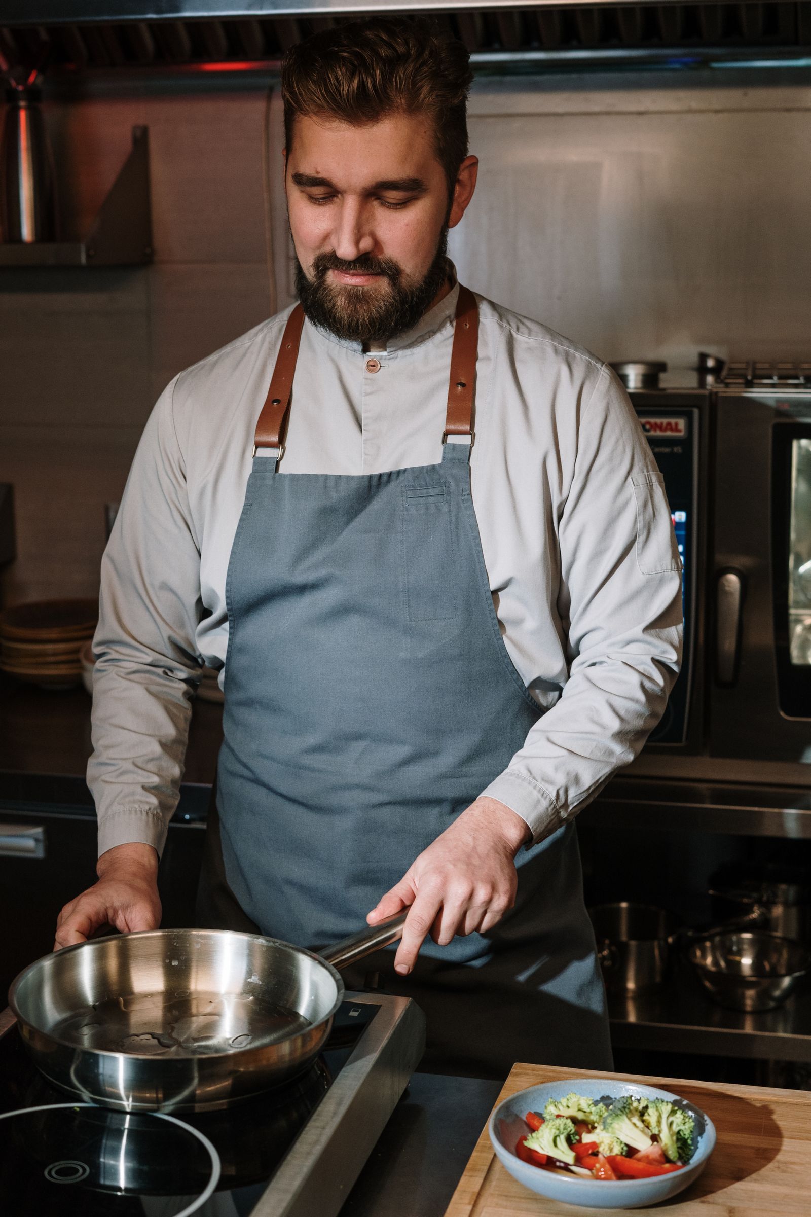 A cook in a long-sleeve shirt and apron heating a pan beside chopped vegetables