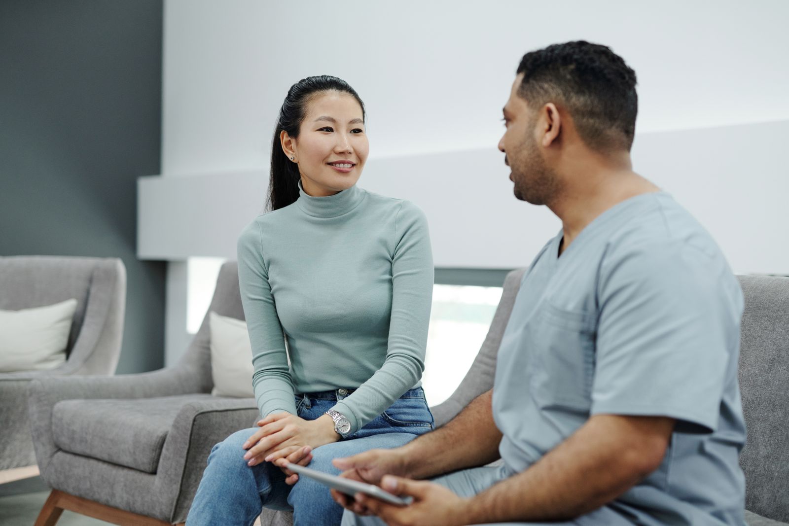 A doctor in clinic clothes talking with a seated patient while holding a tablet