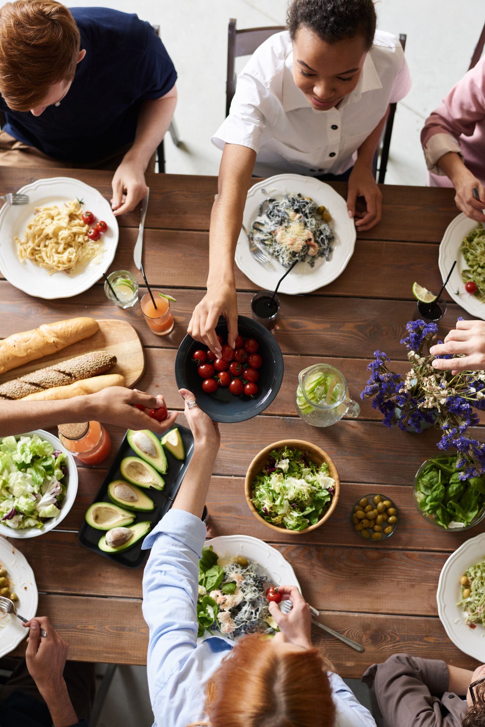 Friends in everyday clothes sharing cherry tomatoes and pasta at a wooden dining table