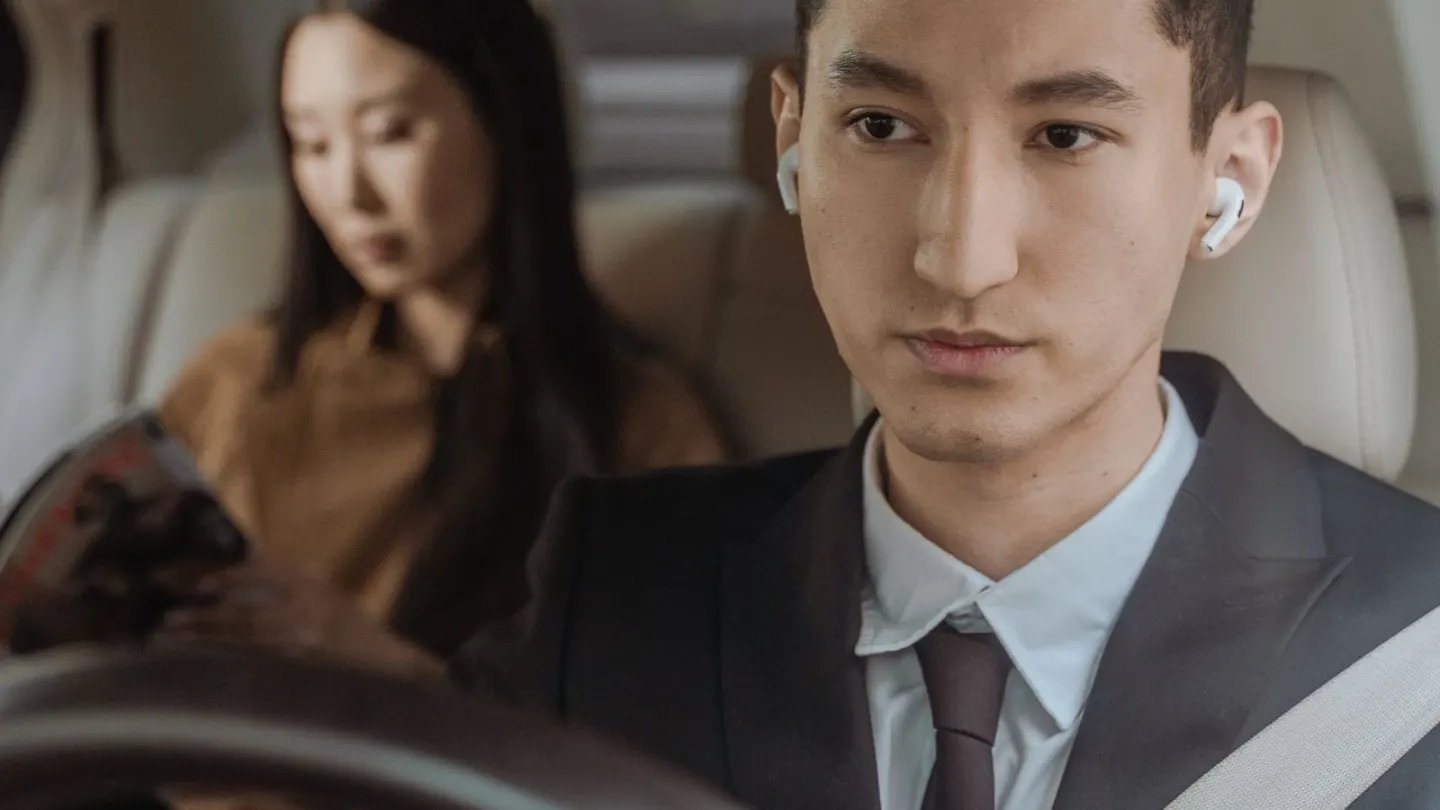 Man in a dark suit and seatbelt driving a car with a passenger in the back