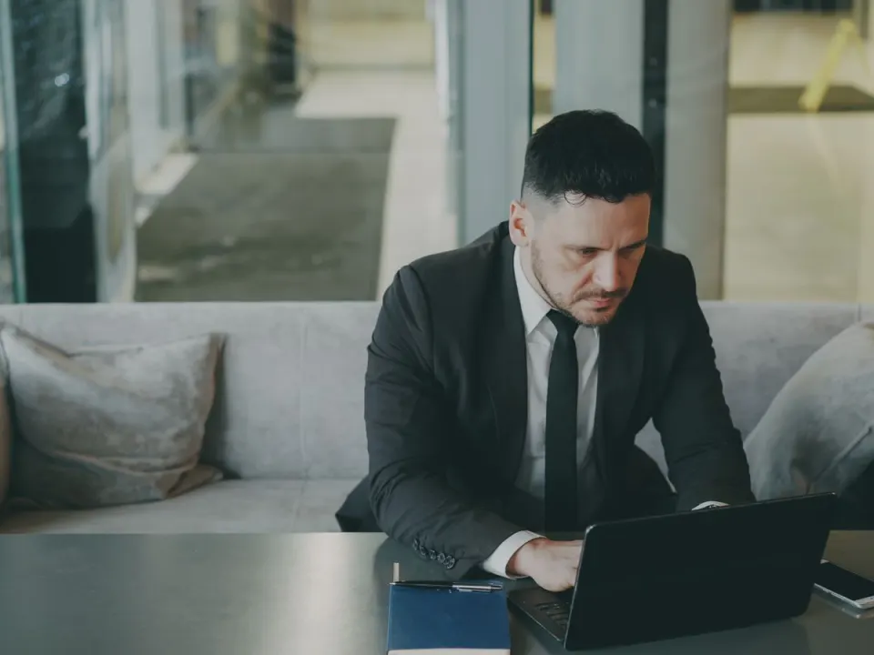 Man in a dark suit working on a laptop at a low table in an office lounge