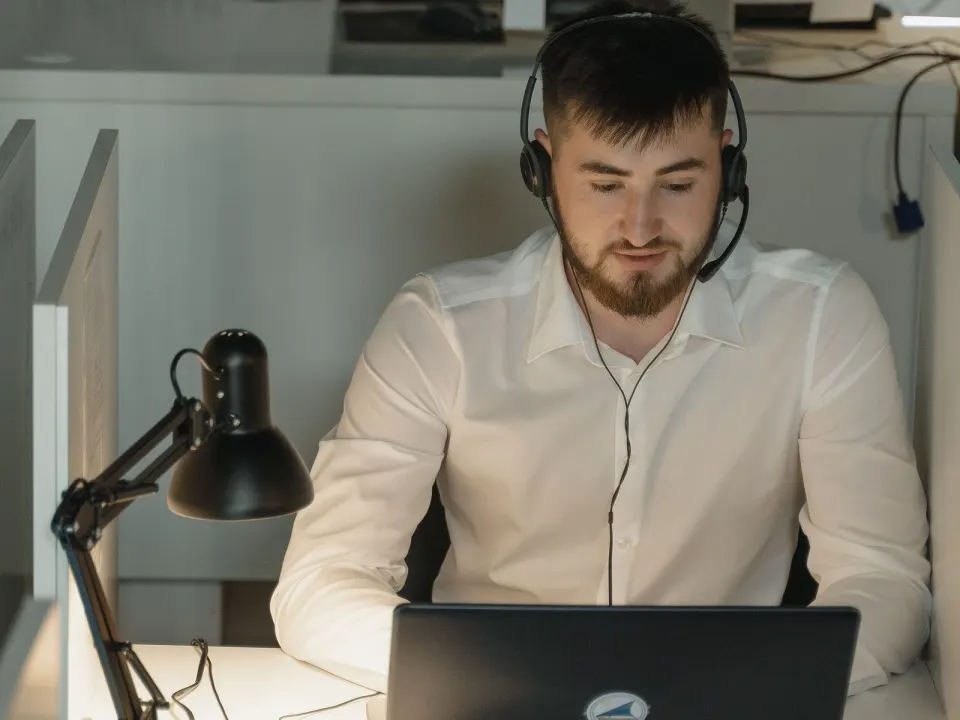 Man in a white shirt wearing a headset while working at a laptop in a desk booth