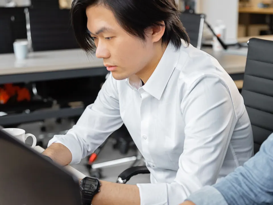 Man in a white shirt working on a laptop at a shared office desk beside a colleague