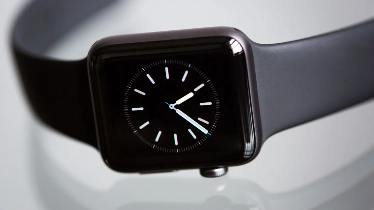 A dark smartwatch showing an analogue clock face on a reflective white surface