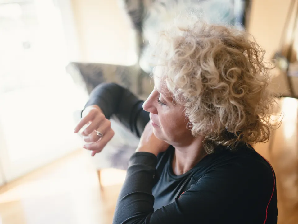Older woman seated on a yoga mat in a bright living room, stretching her neck while checking a watch
