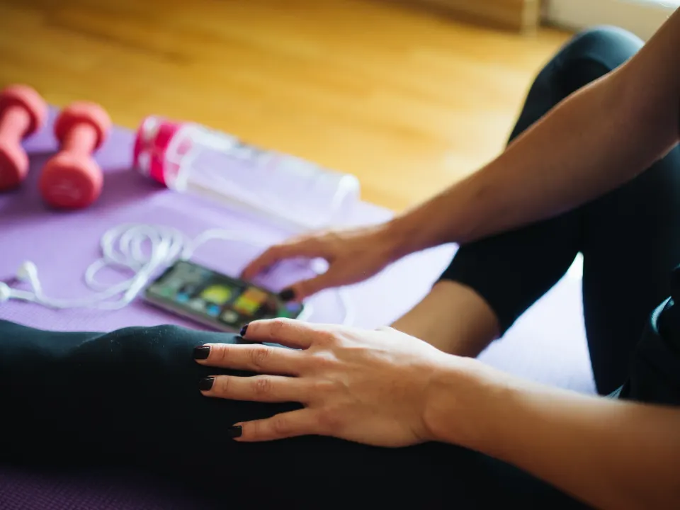 A person in black leggings sits on a purple mat with a phone, water bottle, and small dumbbell nearby