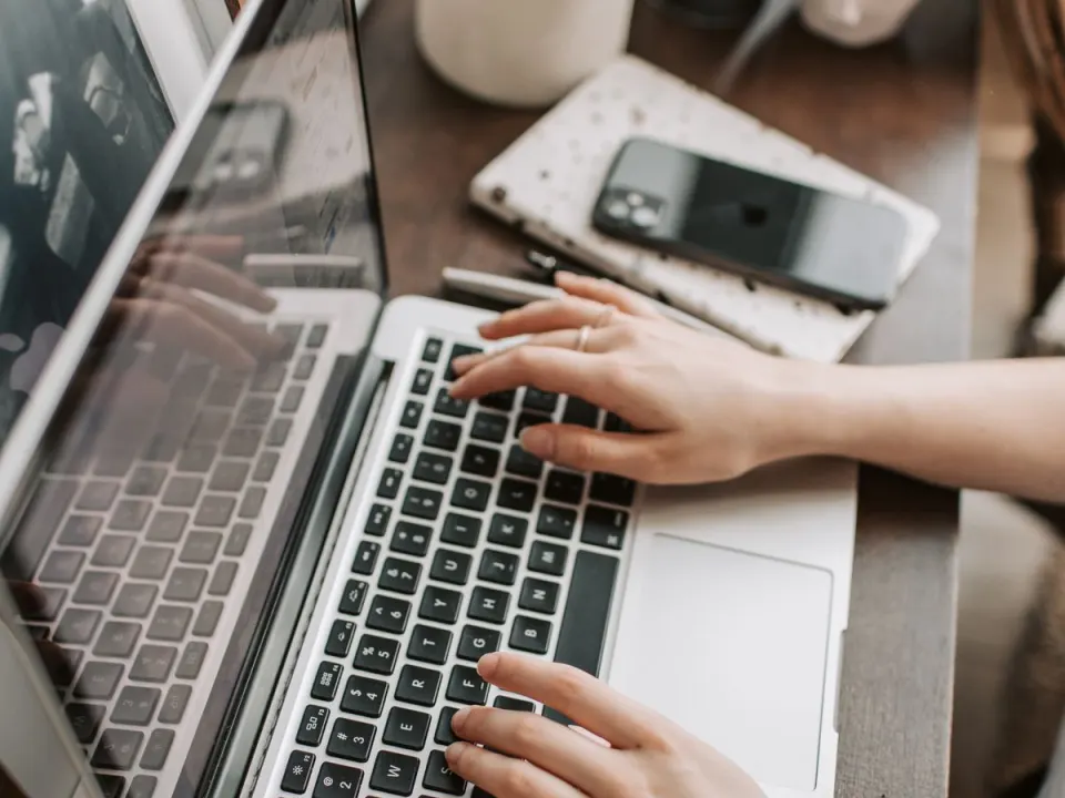 Hands typing on a laptop beside a face-down phone on a wooden desk