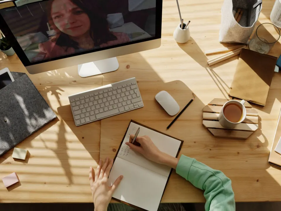 Person in a green jumper writing in a notebook at a sunny wooden desk with tea