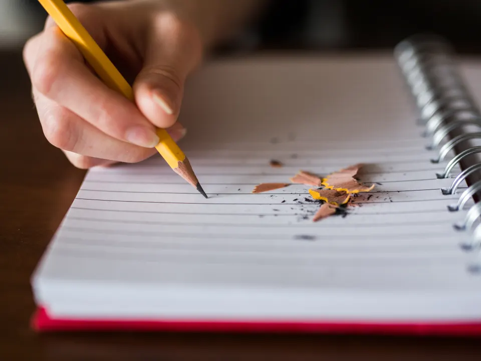A hand holding a yellow pencil above a lined notebook with pencil shavings on the page