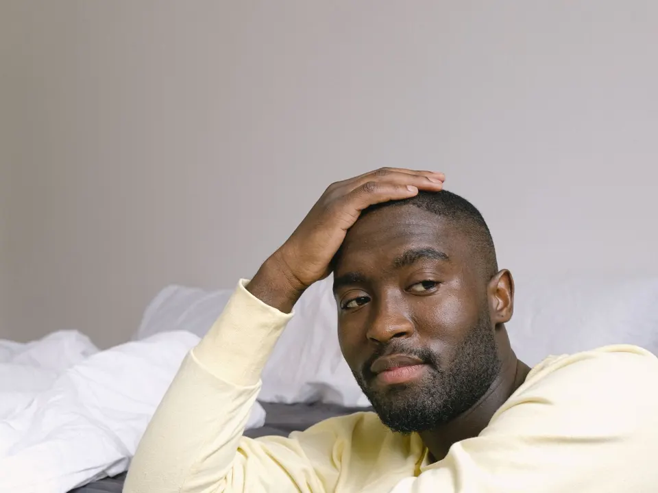 A man in a yellow long-sleeve top sitting on the floor beside a rumpled bed