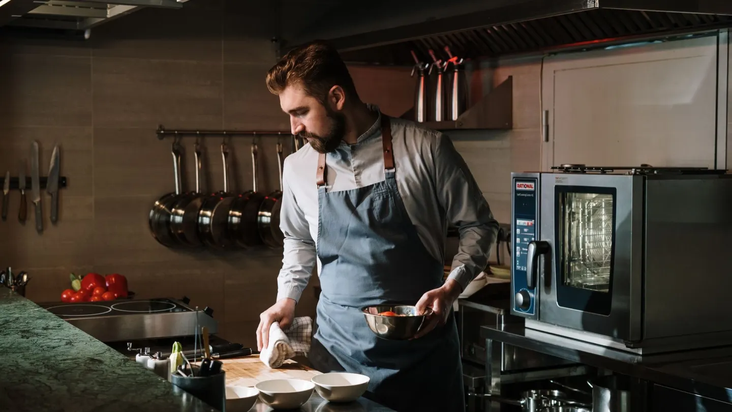 A bearded man in a long-sleeve shirt and blue apron cooking at a kitchen counter