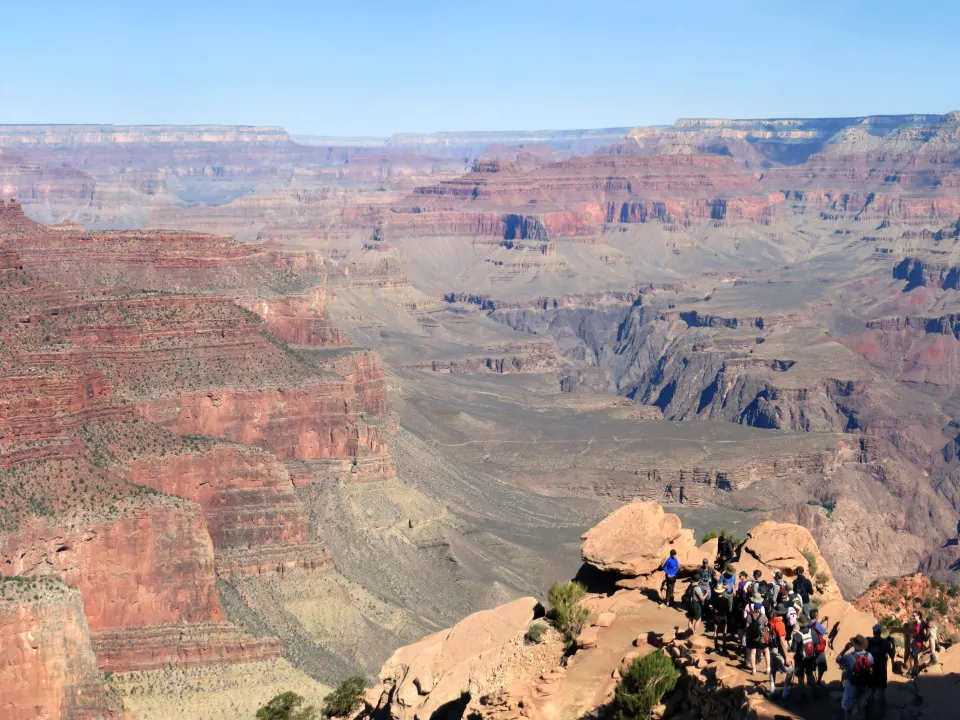 Walkers in modest outdoor clothes stand on a wide desert trail with a canyon view