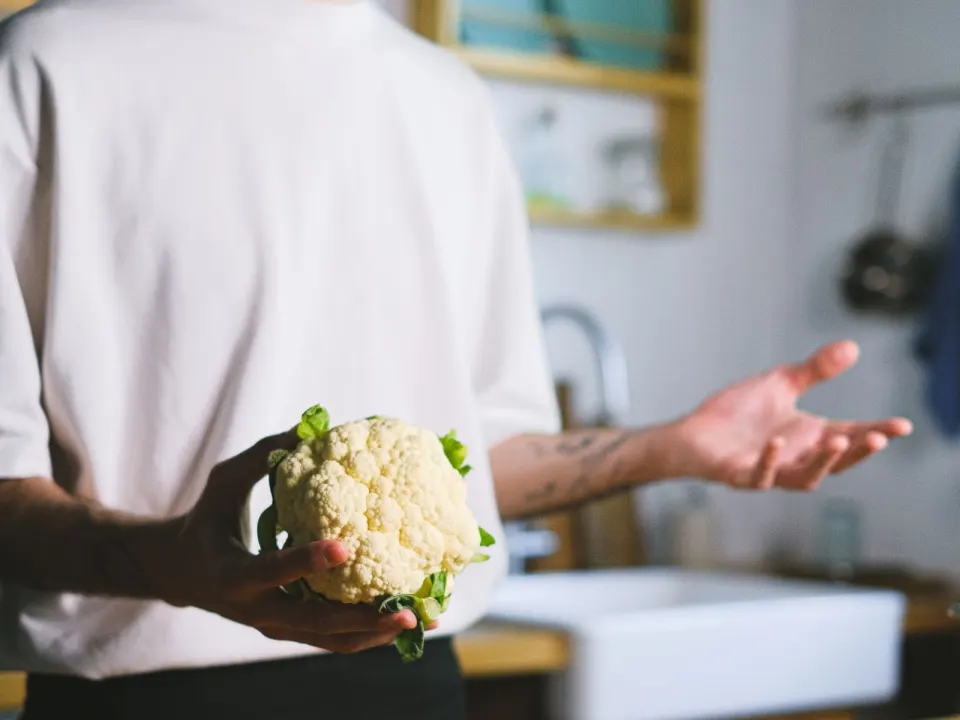 A man in a white T-shirt holding a cauliflower beside tomatoes and peppers