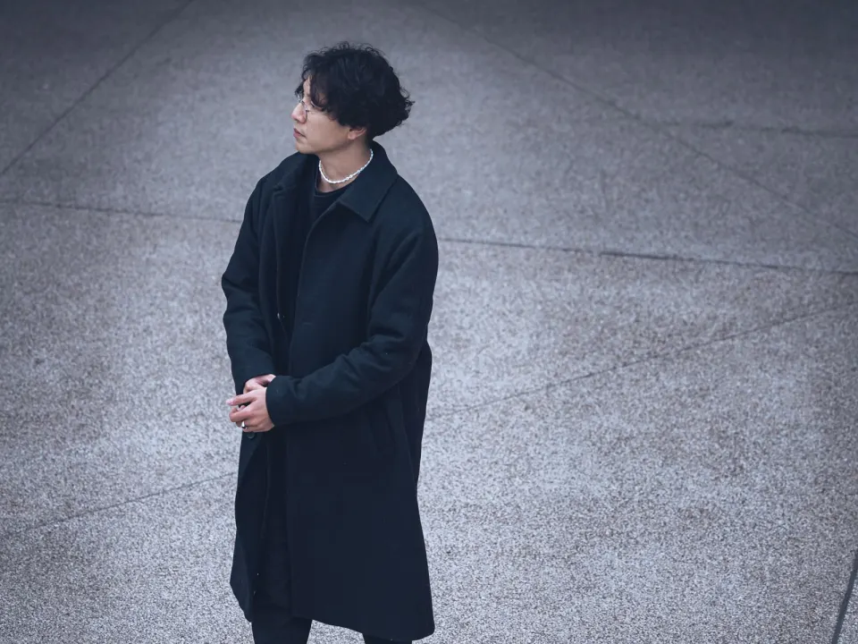 A man in a long dark coat and boots standing on a paved square in daylight