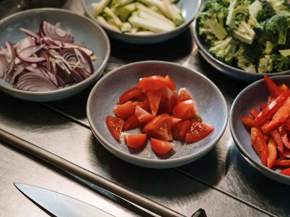 Bowls of chopped tomatoes, peppers, broccoli and onions beside a cook’s knife on a steel counter