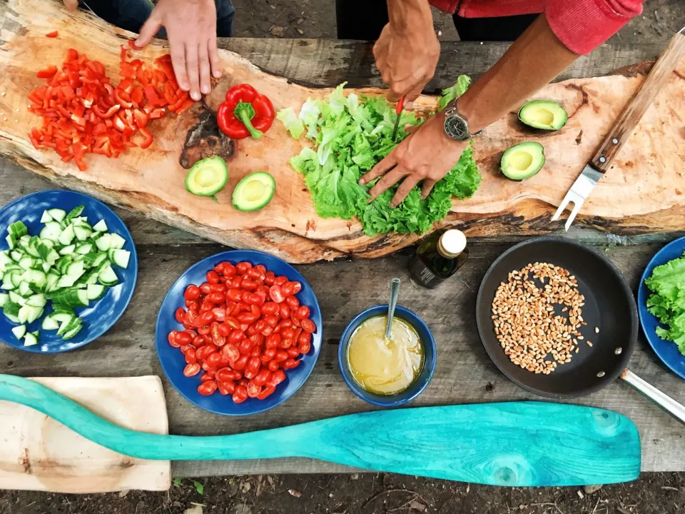 A person in a red jumper chopping lettuce beside bowls of tomatoes, cucumber, and peppers on a wooden table