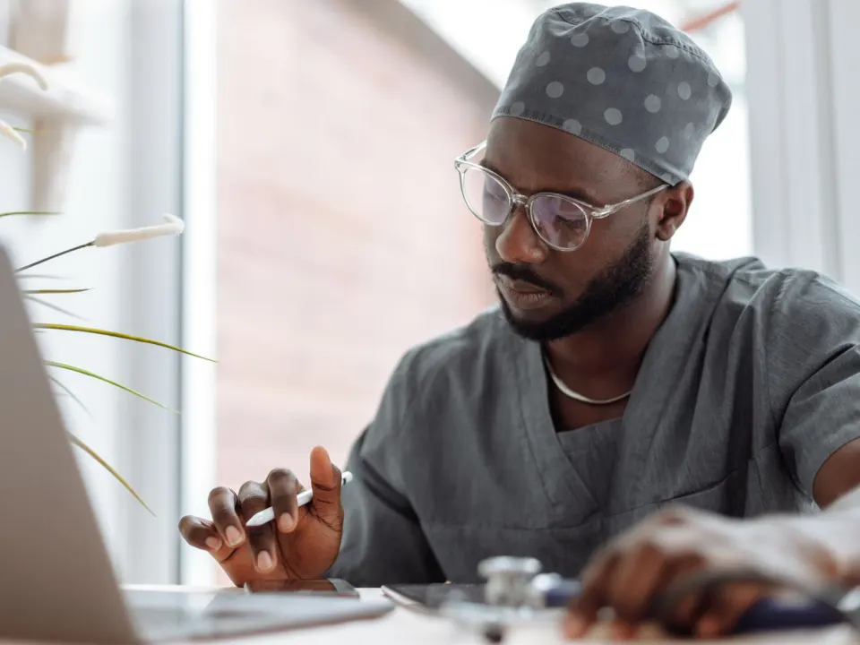 A clinician in grey scrubs and a cap reviewing notes on a laptop at a desk
