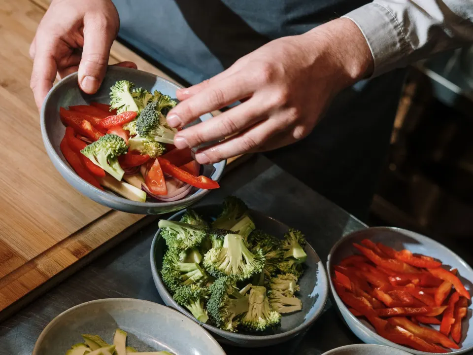 A cook in a shirt and apron arranging bowls of chopped peppers, broccoli and tomato