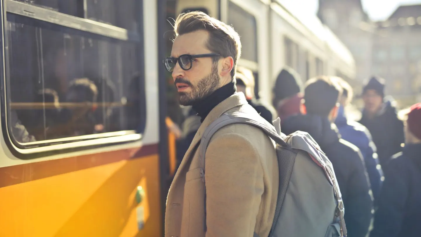A bearded man in a beige coat and glasses waiting beside a tram with a backpack