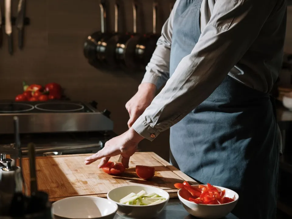 A cook in a long-sleeved shirt and apron slicing tomatoes on a wooden board