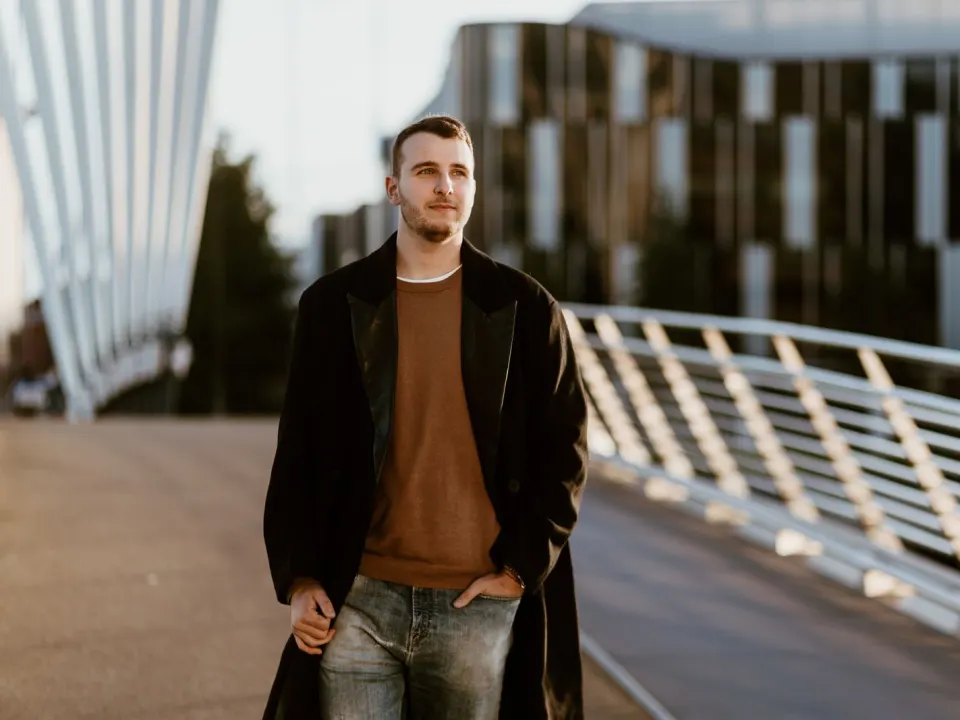 A man in a brown jumper and dark coat walking on a city footbridge in daylight