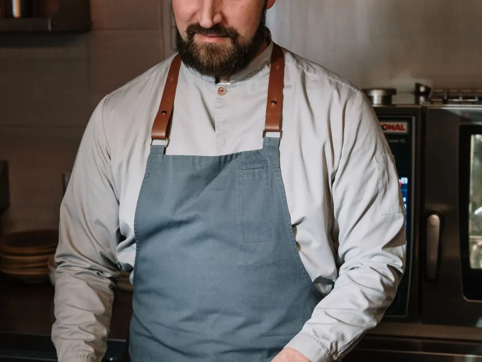 A cook in a long-sleeve shirt and apron heating a pan beside chopped vegetables