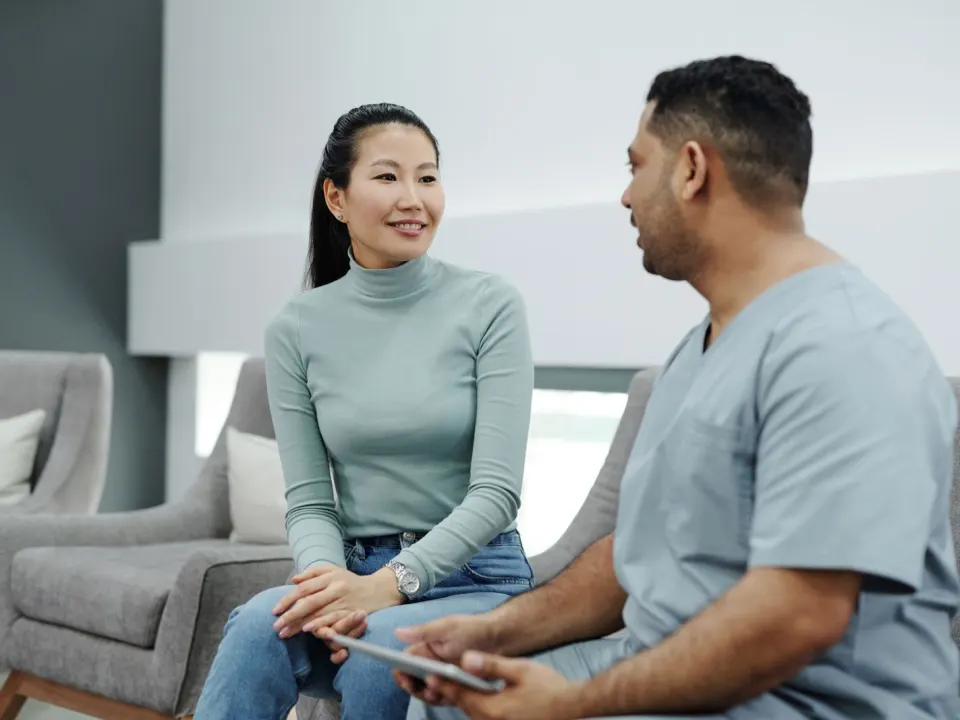 A doctor in clinic clothes talking with a seated patient while holding a tablet