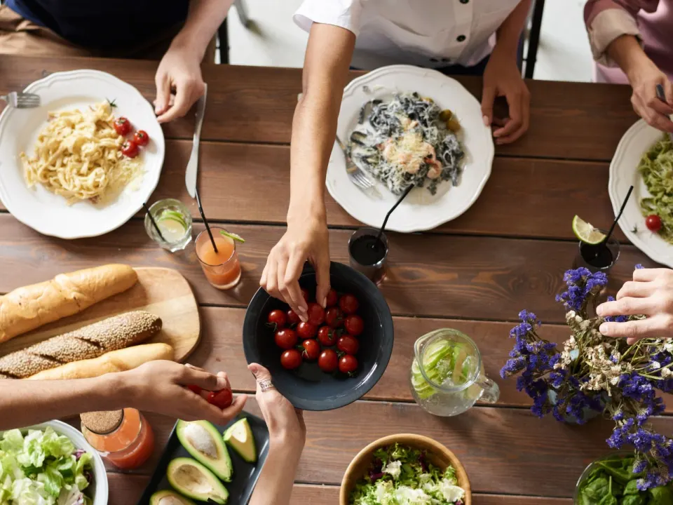 Friends in everyday clothes sharing cherry tomatoes and pasta at a wooden dining table